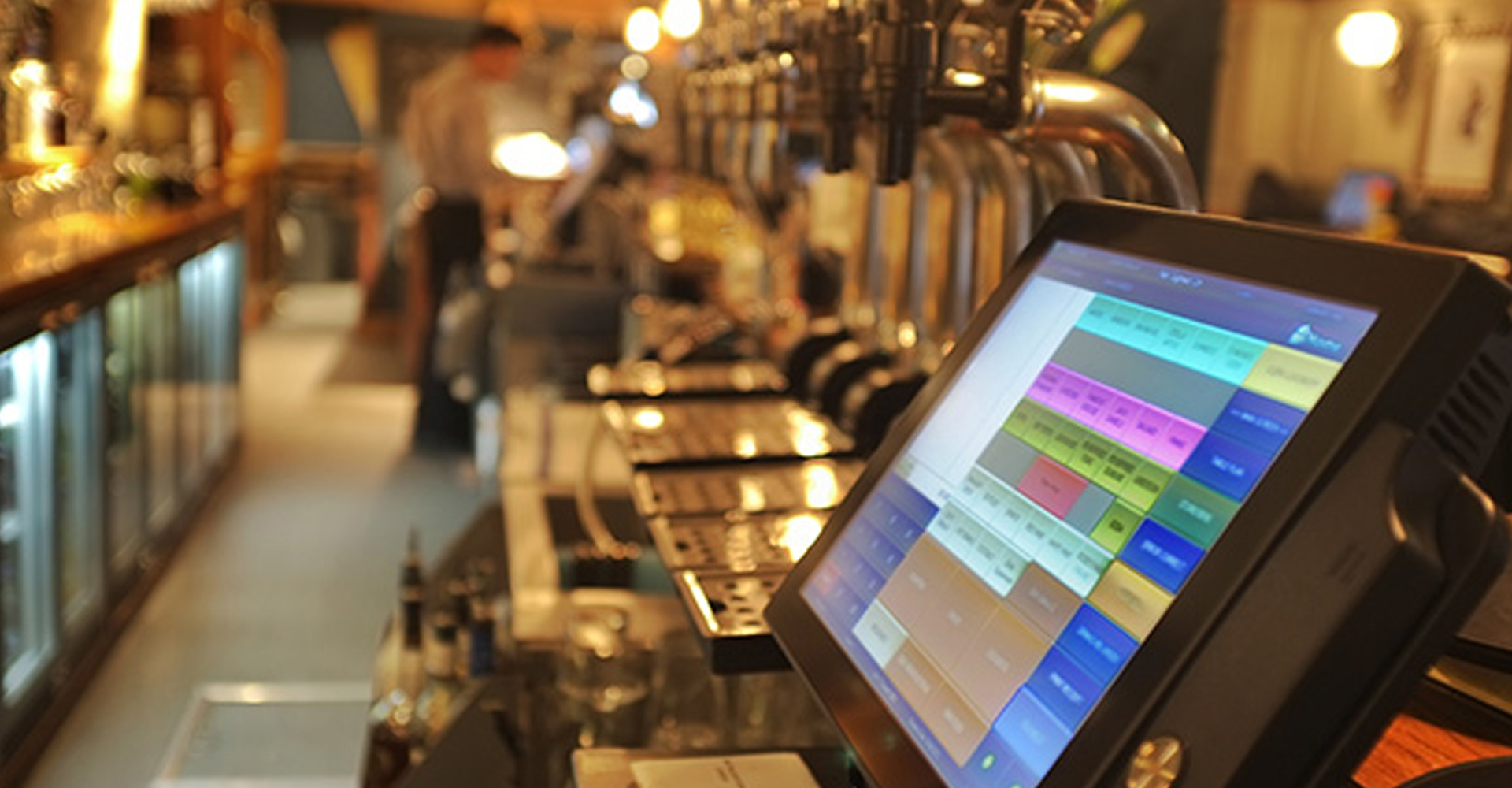 Point of sale touchscreen system by a bar counter with multiple beer taps in a dimly lit pub or bar.