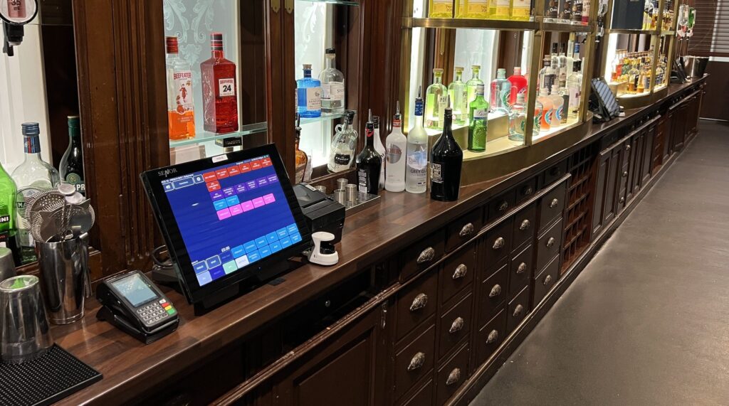 Long wooden bar counter with a variety of liquor bottles displayed along the back, a touchscreen point-of-sale system, and a card payment terminal on the counter.