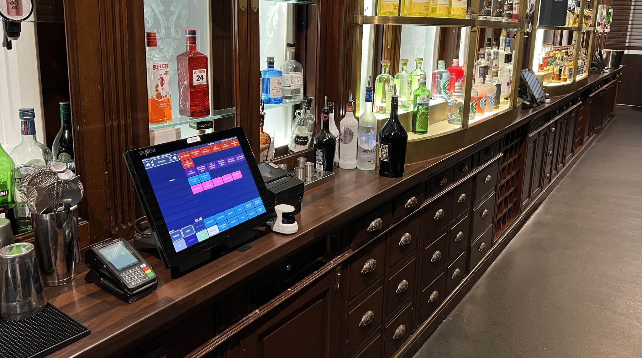 Long wooden bar counter with a variety of liquor bottles displayed along the back, a touchscreen point-of-sale system, and a card payment terminal on the counter.