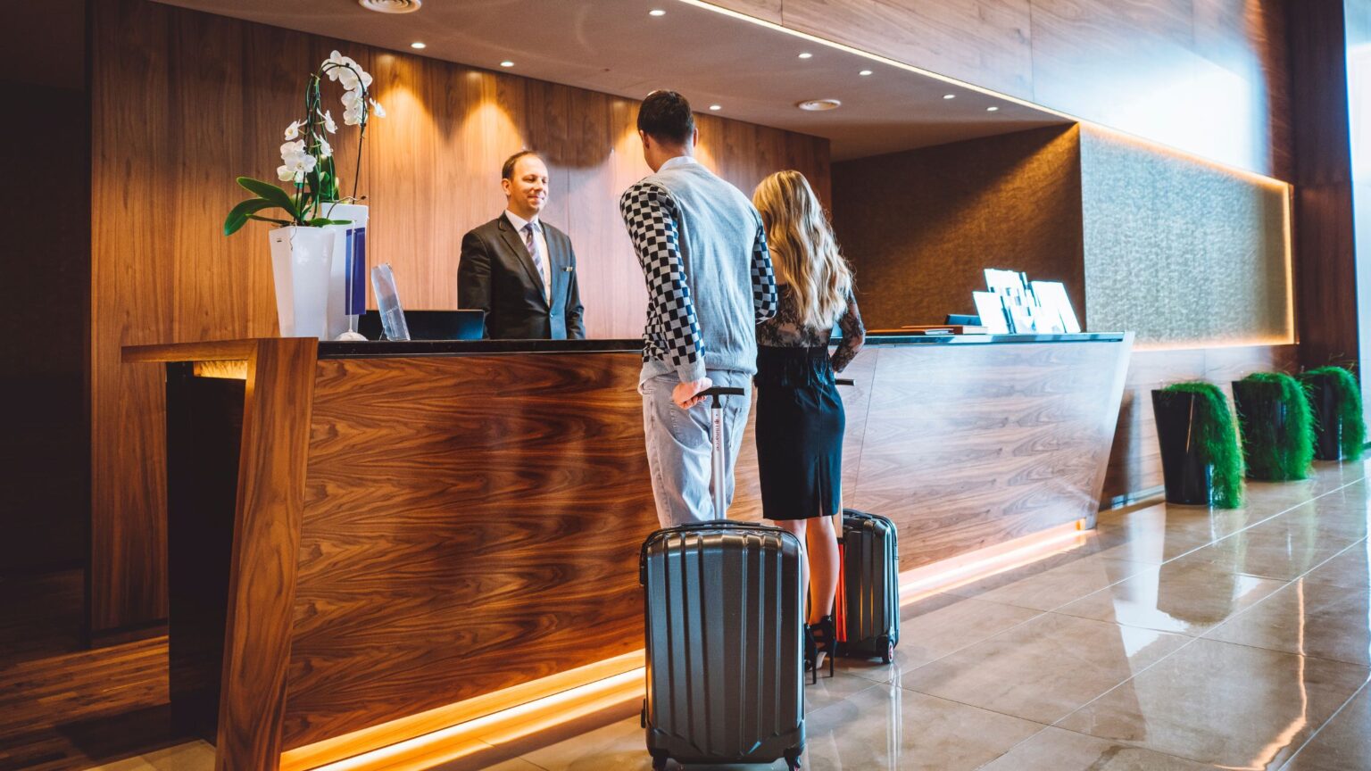 A couple with suitcases checks in at a modern hotel reception desk, speaking with a suited receptionist.